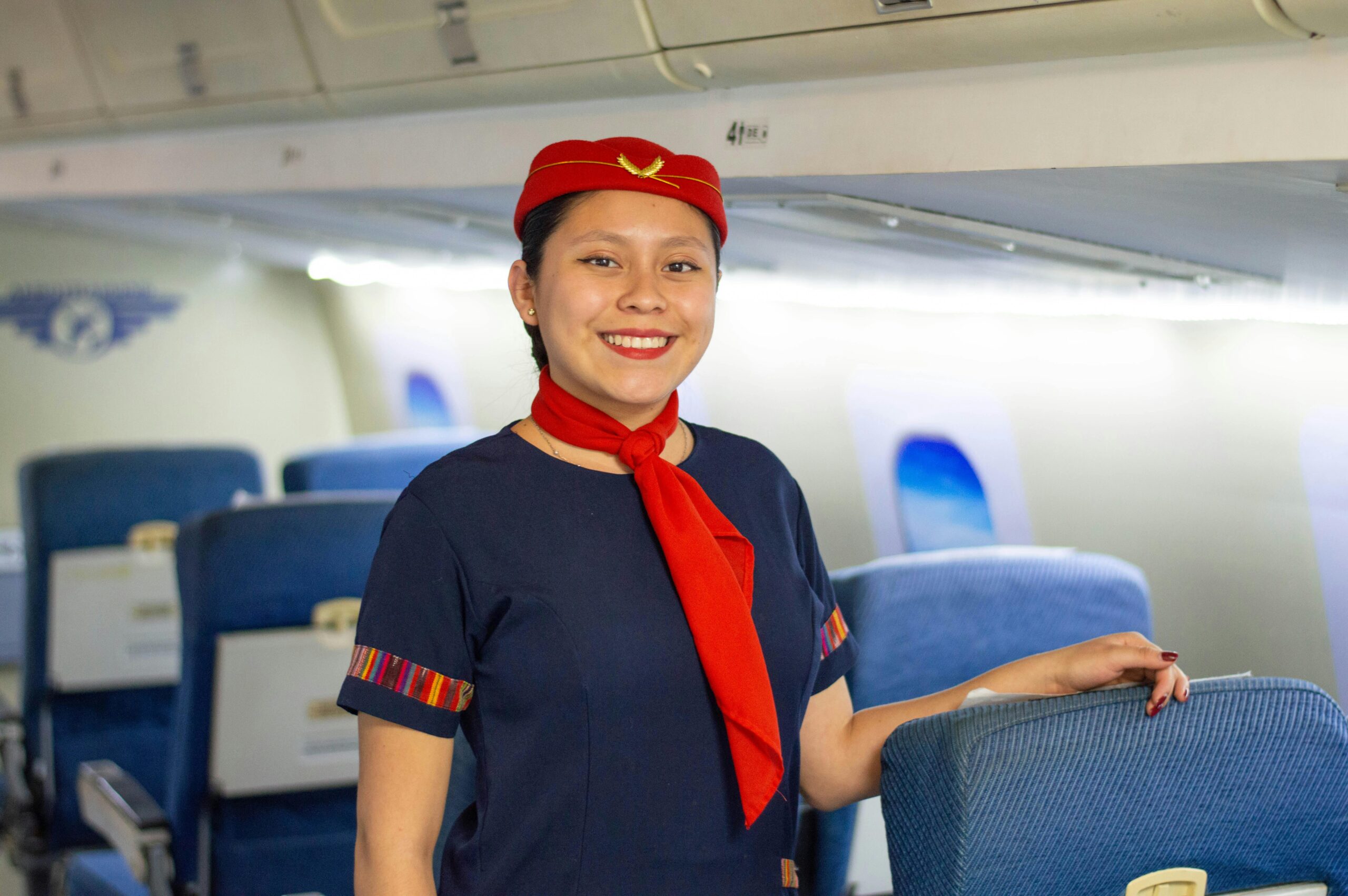 Enthusiastic flight attendant wearing a red uniform stands smiling in an airplane cabin.